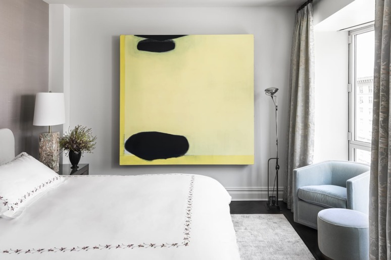 Bright modern bedroom with large abstract yellow and black painting, white linens, light blue chair, and tall window.