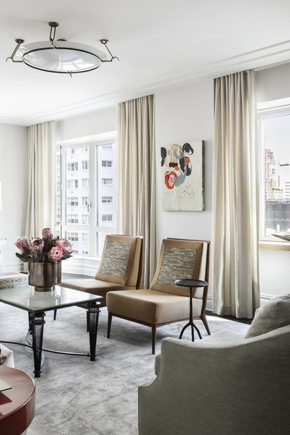 Modern living room with beige chairs, a glass coffee table, floral arrangement, art on the wall, and large windows with curtains.