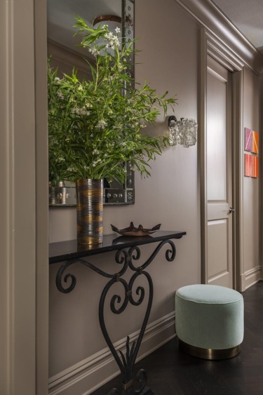 Elegant hallway with decorative console table, green plant in vase, wall mirror, artwork, and a teal ottoman.