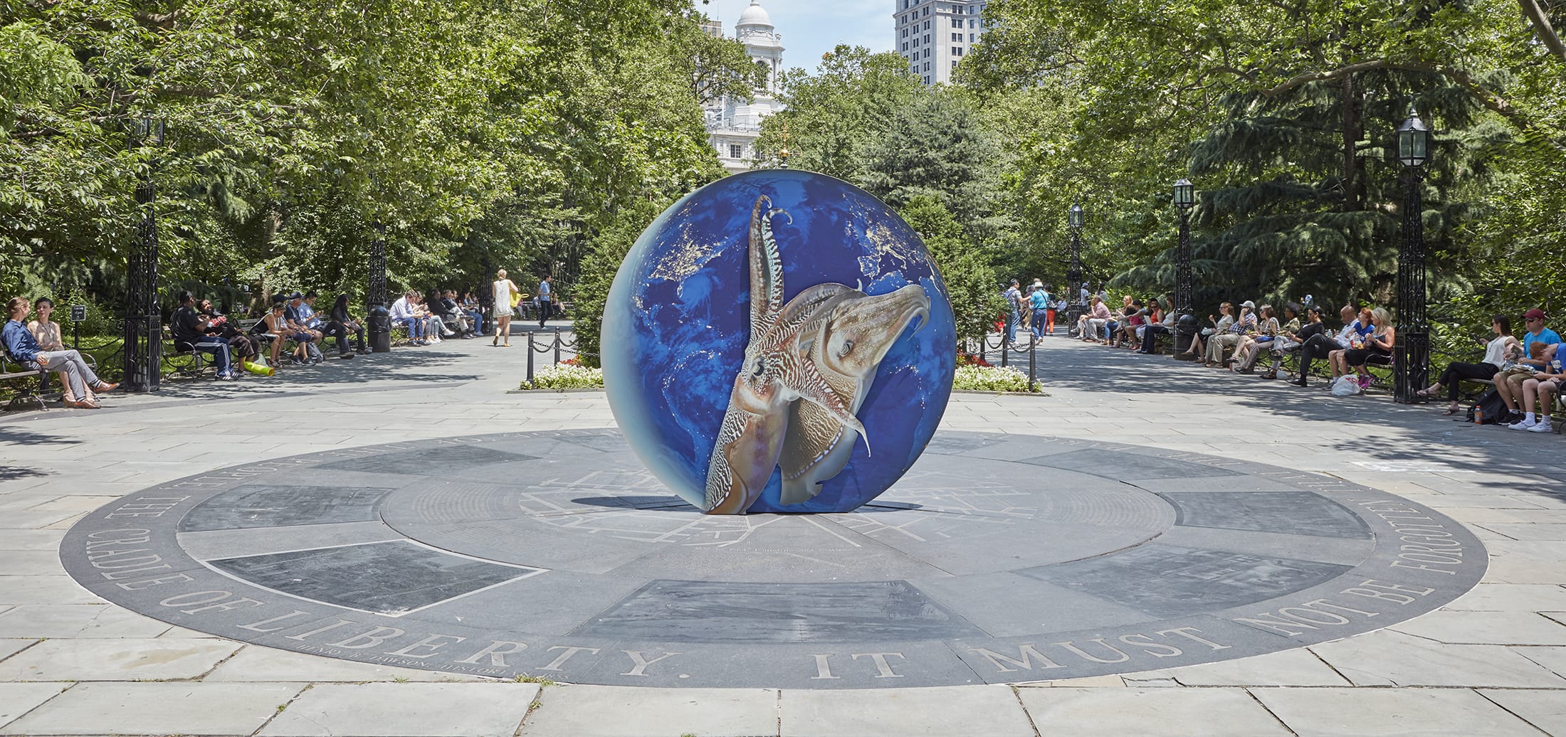 Shark sculpture displayed outdoors on a circular stone platform surrounded by trees and people sitting on benches.