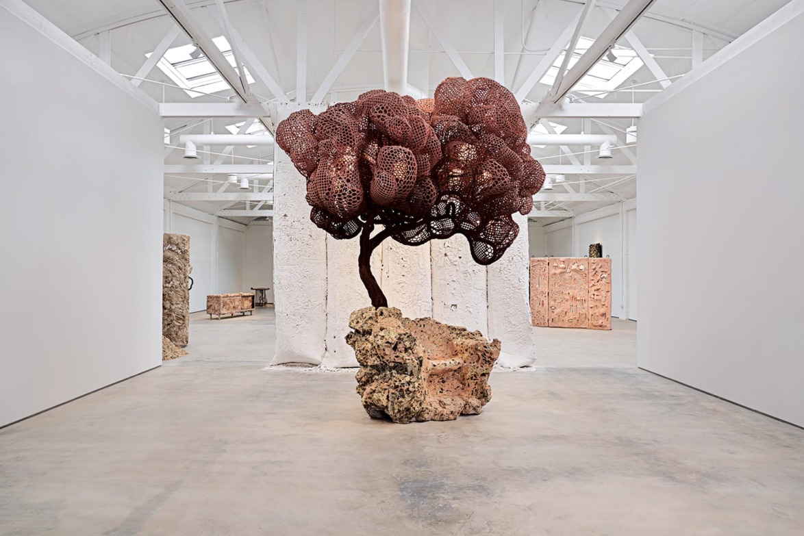 Sculpture of organic red structure atop a textured stone base in a spacious white gallery with natural light.