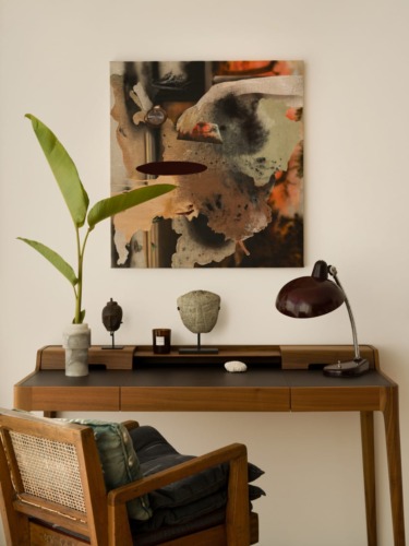 Cozy workspace with wooden desk, artistic abstract wall art, potted plant, and chair in neutral-toned room.