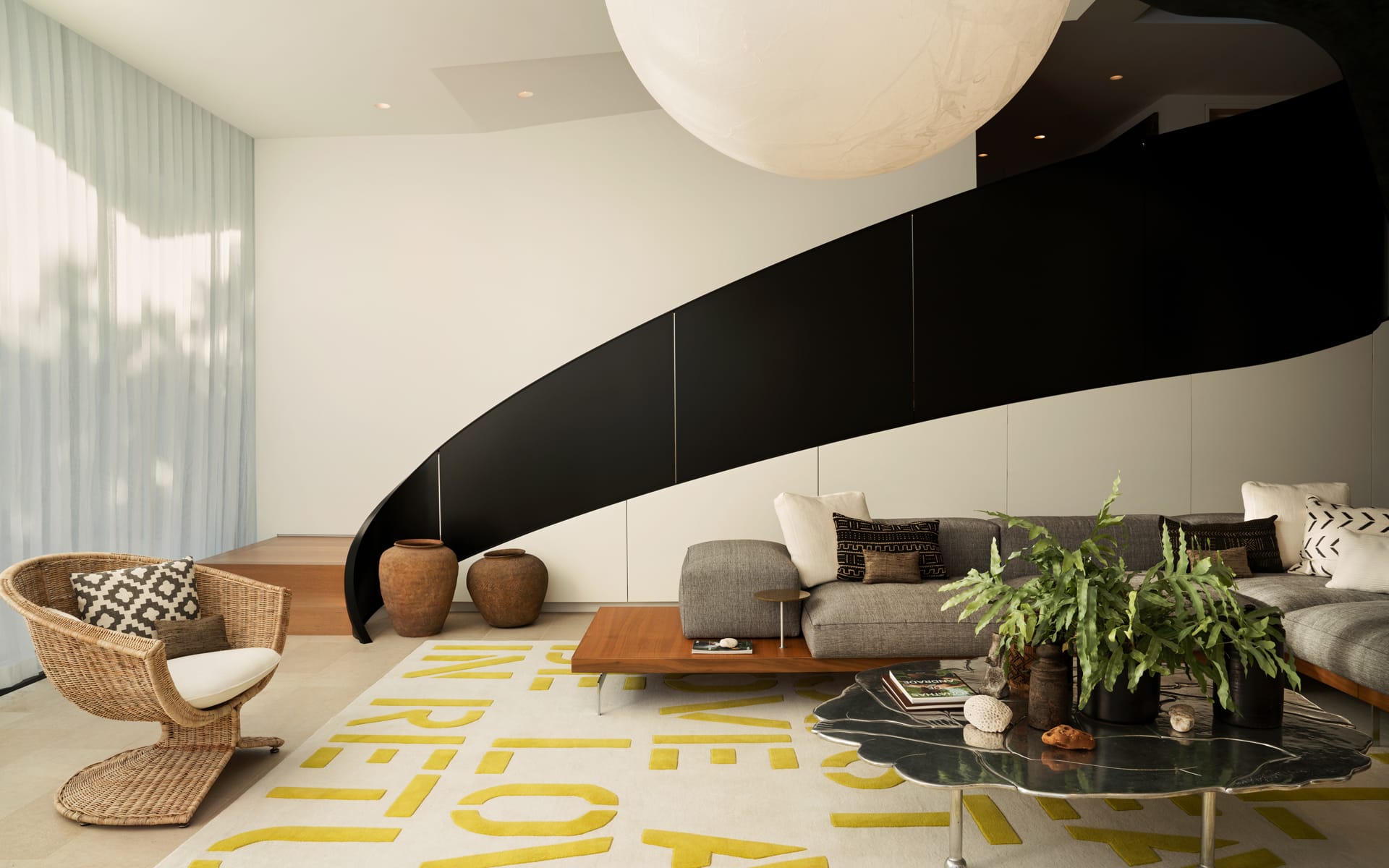 Modern living room with curved black staircase, stylish furniture, green plant, woven chair, and large spherical light fixture.