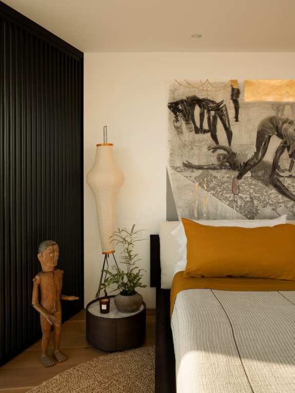 Cozy bedroom with abstract art, wooden sculpture, and a tall lamp beside a neatly made bed with mustard and white linens.