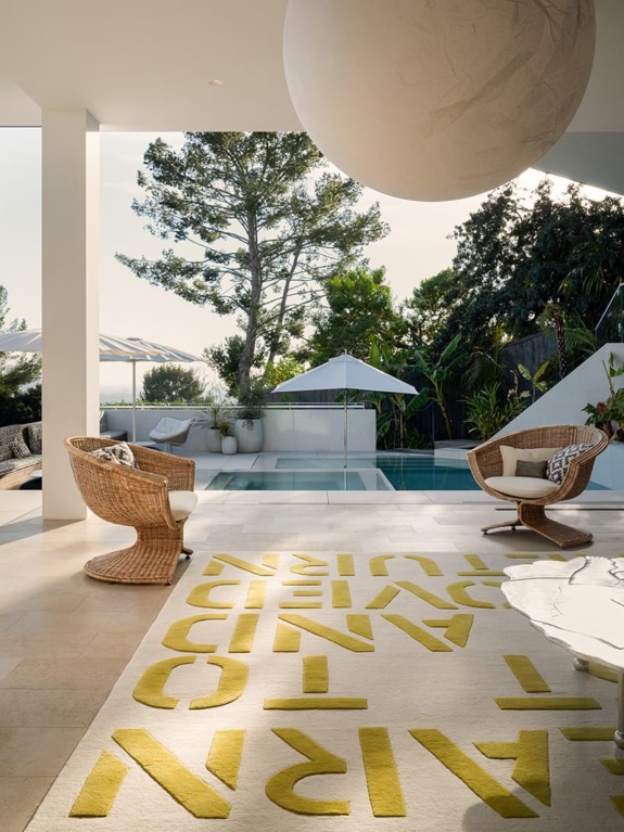 Outdoor seating area with wicker chairs and modern design, overlooking a pool and garden with trees and umbrellas.