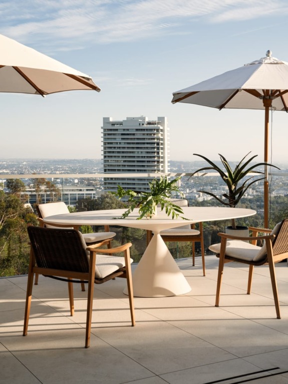 Outdoor patio with a round table, chairs, potted plants, and umbrellas overlooking cityscape and tall white building in background.