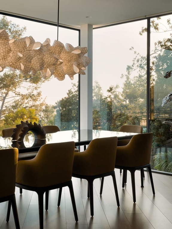 Modern dining room with large windows, tree view, yellow chairs around a glass table, and a unique artistic chandelier.