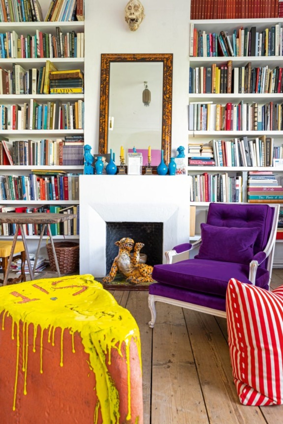 Colorful living room with purple chair, bookshelves, decorative fireplace, and striped coffee table in eclectic design.