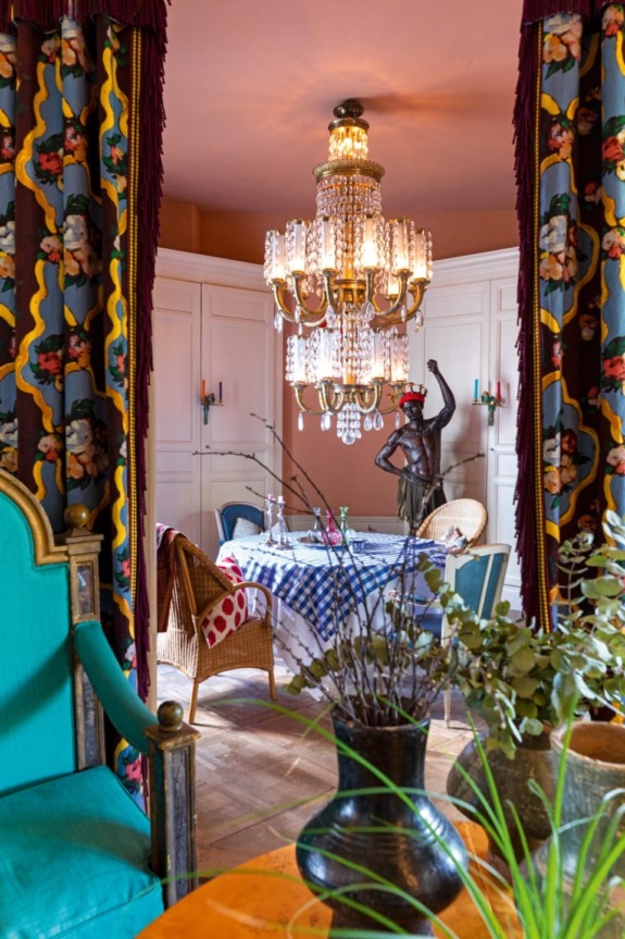 Eclectic dining room with bright colors, a chandelier, patterned curtains, and various plants and sculptures.