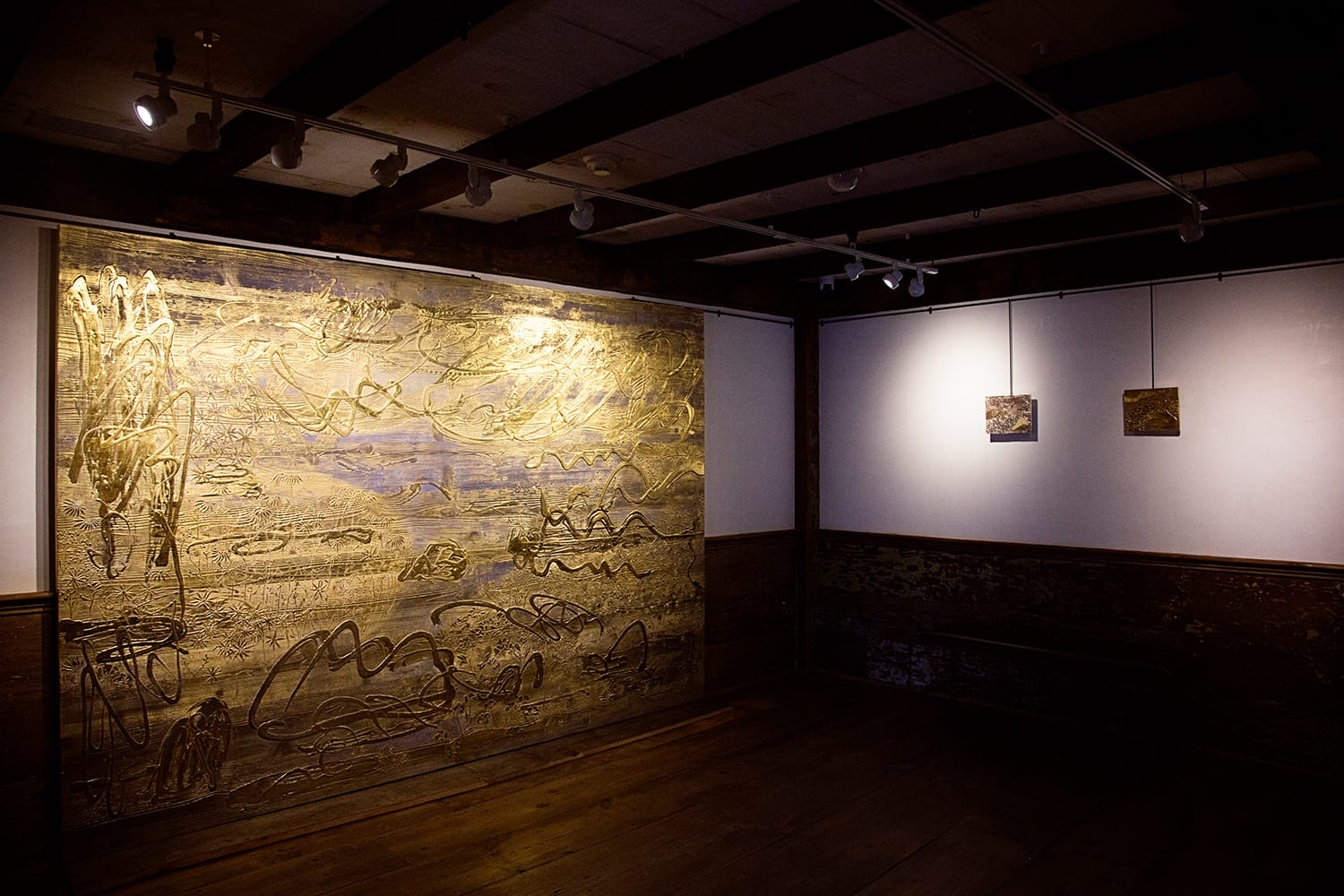Gallery room with large abstract artwork on the left wall, featuring gold tones and dynamic patterns, small paintings on the right.