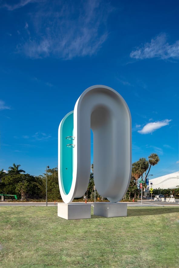 Large modern sculpture with a curved shape, featuring a turquoise interior, set against a clear blue sky and grassy area.