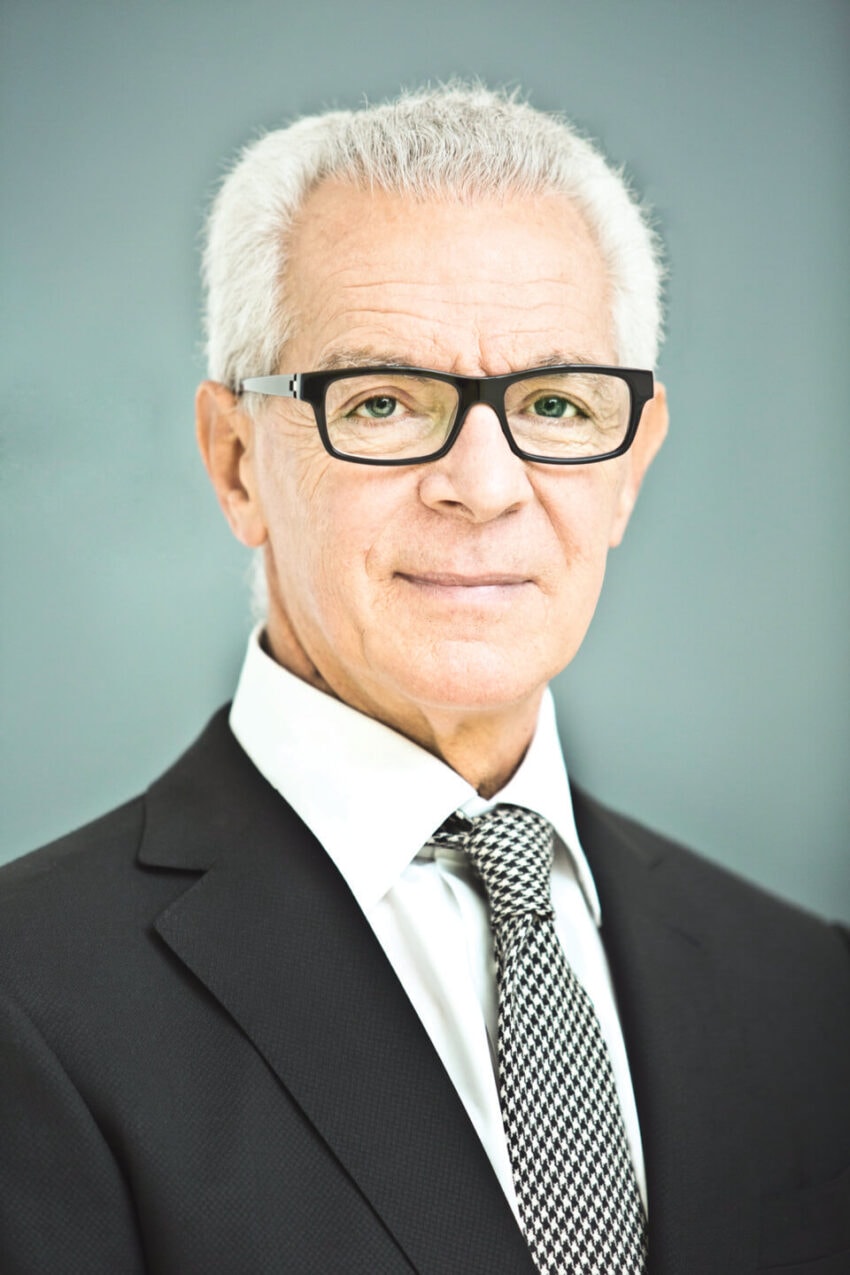 Elderly man with glasses wearing a suit and tie against a plain background
