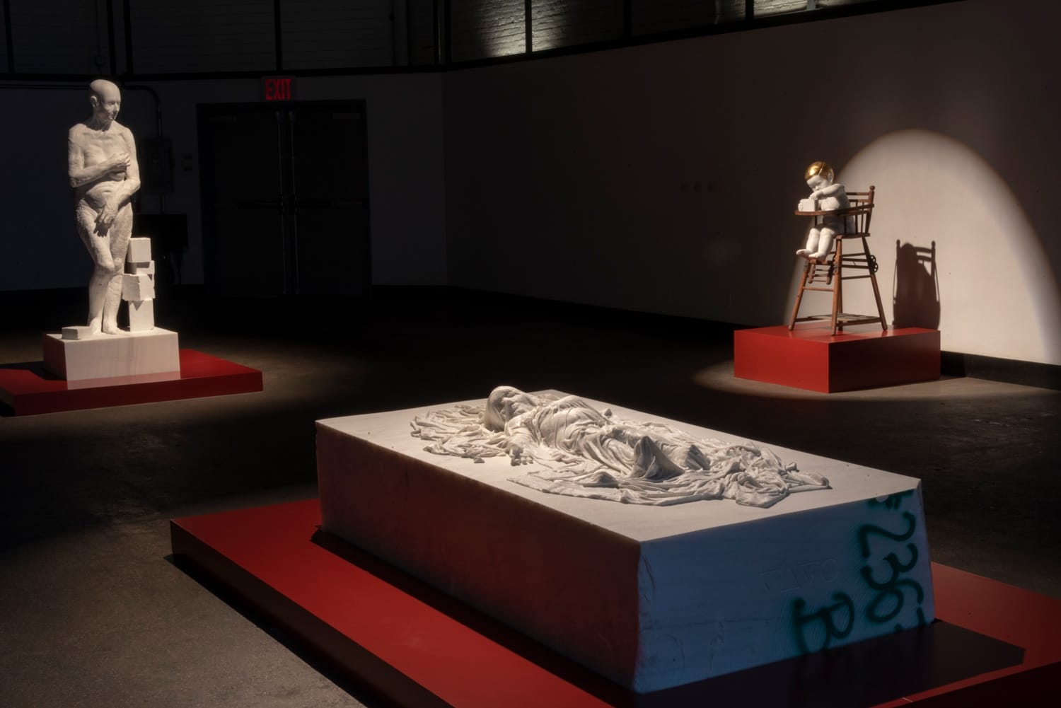 Art exhibition with three sculptures on red platforms, including a reclining figure and a child on a chair, in dim lighting.