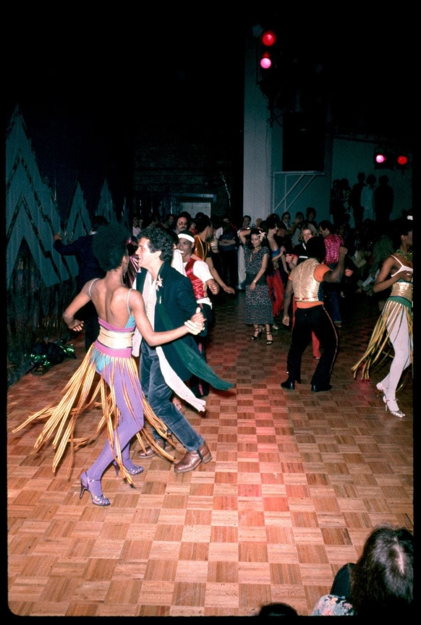 People dancing energetically in a lively club setting with colorful outfits and a wooden dance floor.