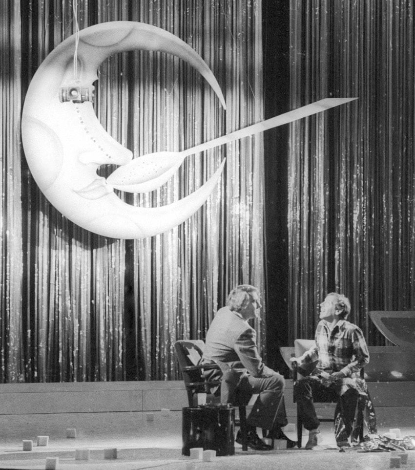 Two people sit on stage with a crescent moon prop, surrounded by curtain-like backdrop and glittering streamers.