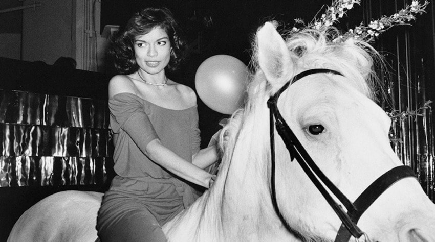 A person riding a white horse at an indoor event, holding a balloon, with decorations in the background.