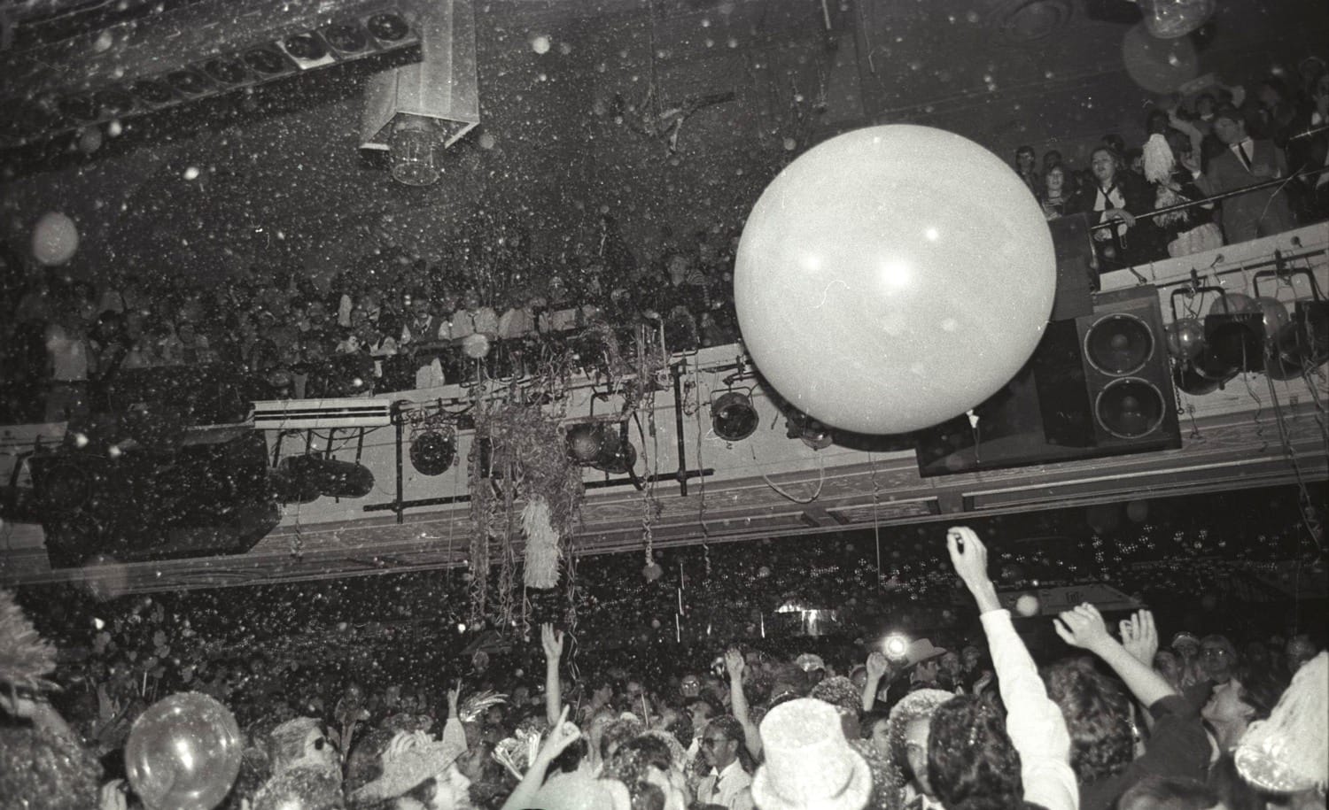 Crowded party scene with people dancing, large balloon in air, and confetti falling in a festive, energetic atmosphere.