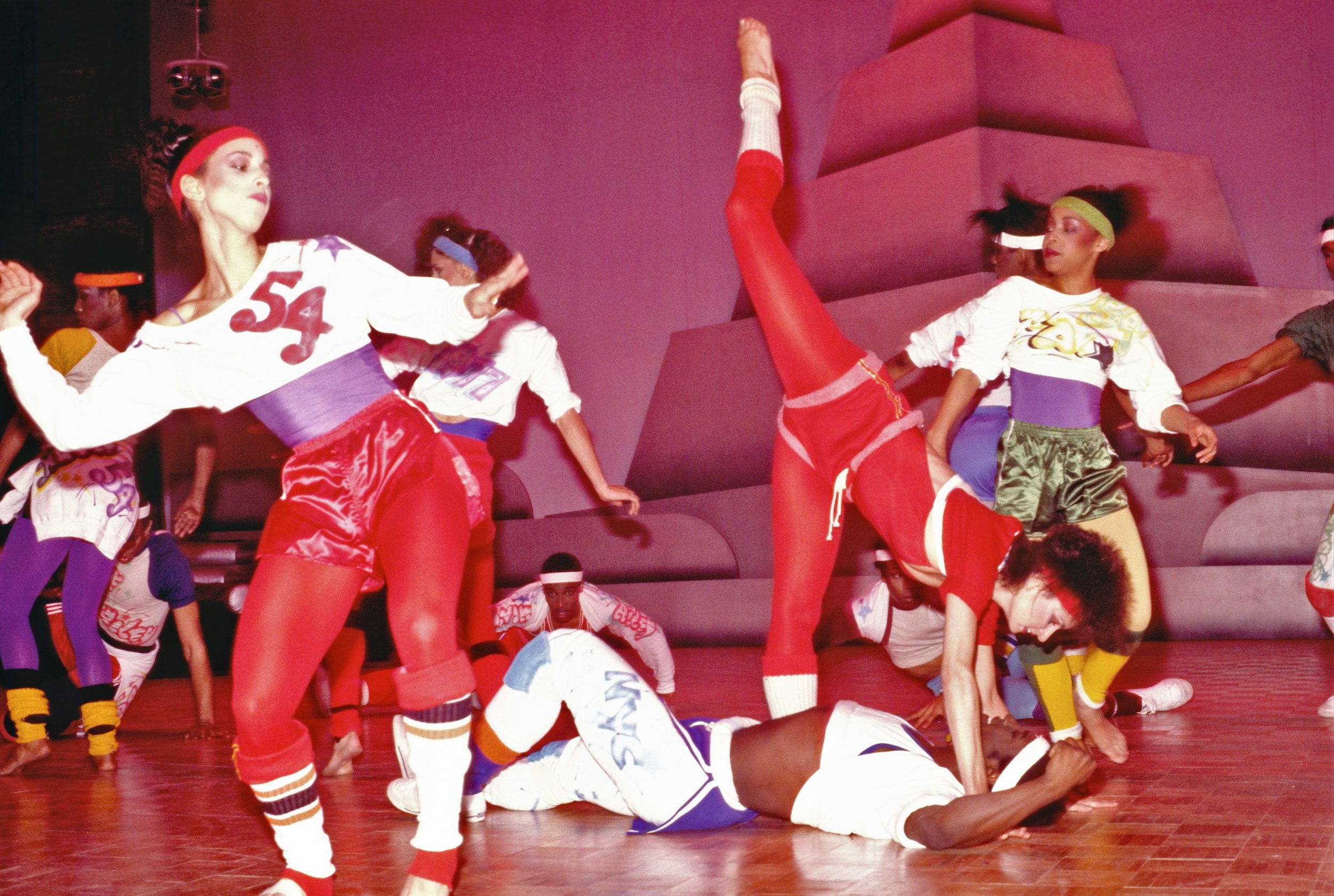 Dancers in colorful 1980s athletic wear performing energetically on a stage with a geometric backdrop.