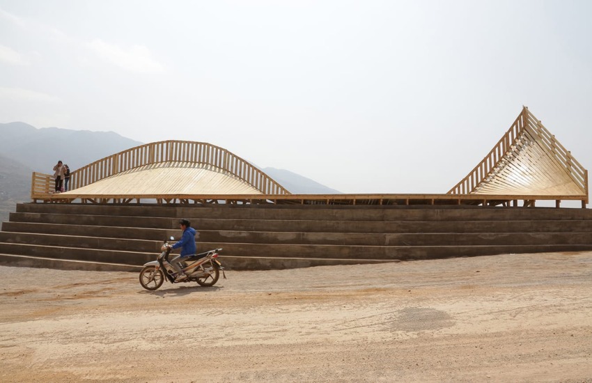 Person on motorcycle rides past a unique wooden architectural structure with people walking on it and mountains in the background.