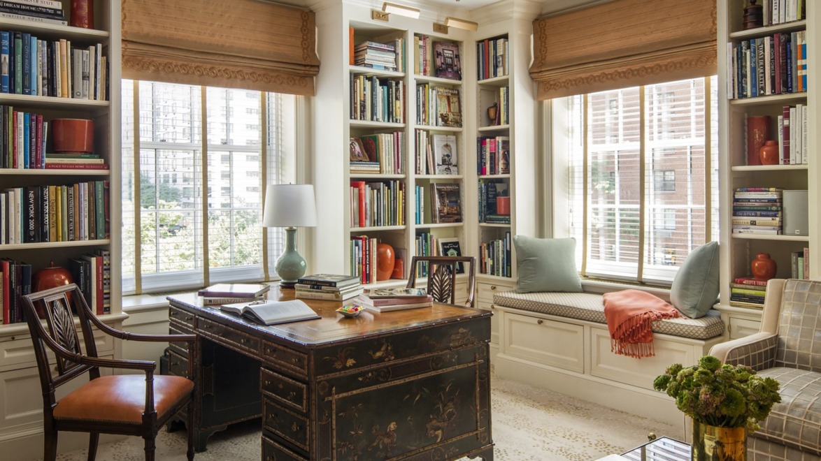Elegant home office with a vintage desk, wooden chair, bookshelves, large windows, and cozy window seating with cushions.