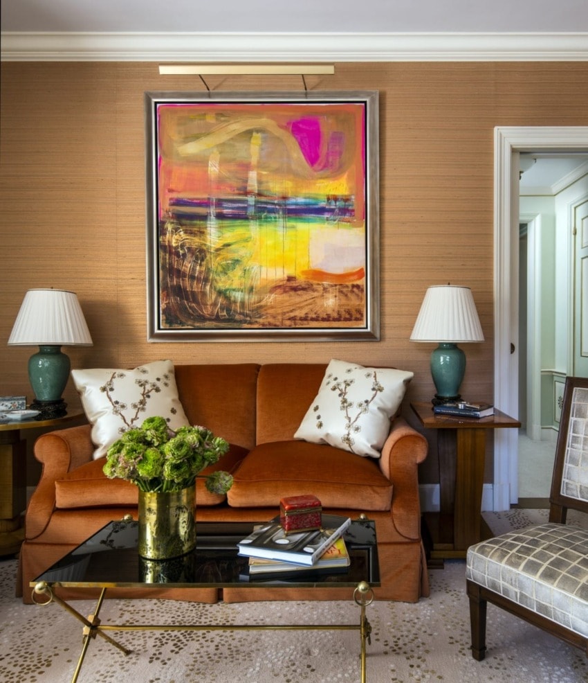 Orange sofa with decorative pillows, abstract art above, green lamps on wooden side tables, and a glass coffee table with flowers.