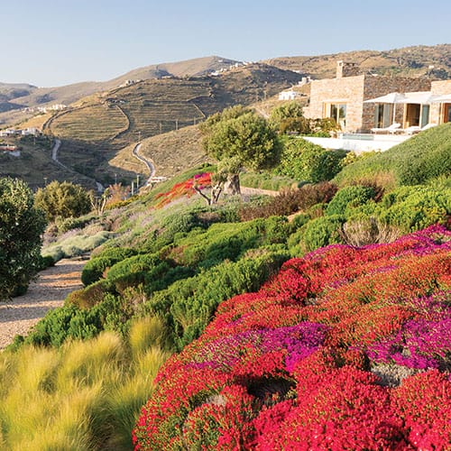 Landscape view of vibrant gardens with colorful flowers and lush greenery overlooking rolling hills and a stone building.
