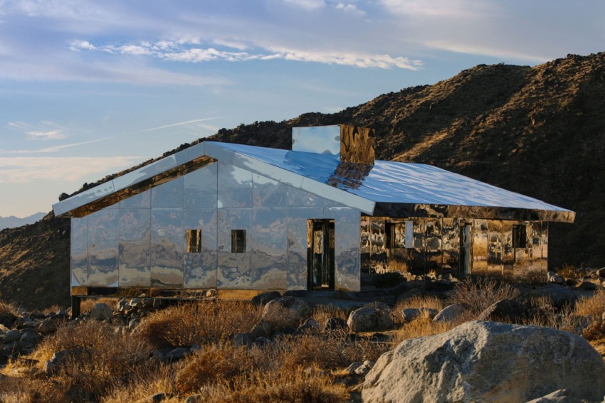 Desert landscape with a modern, reflective glass house surrounded by rocks and hills under a partly cloudy sky.