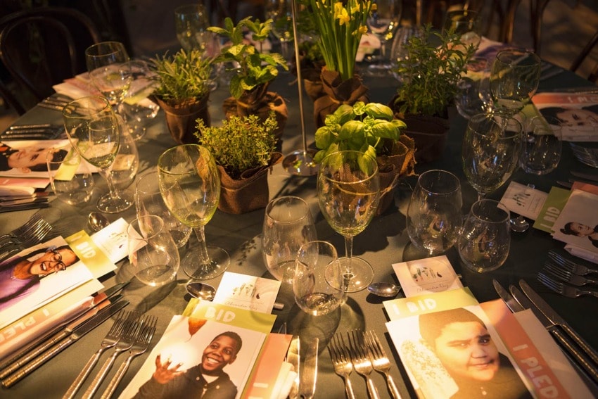 Elegant dinner table setting with place cards, wine glasses, cutlery, and small potted plants as centerpieces.
