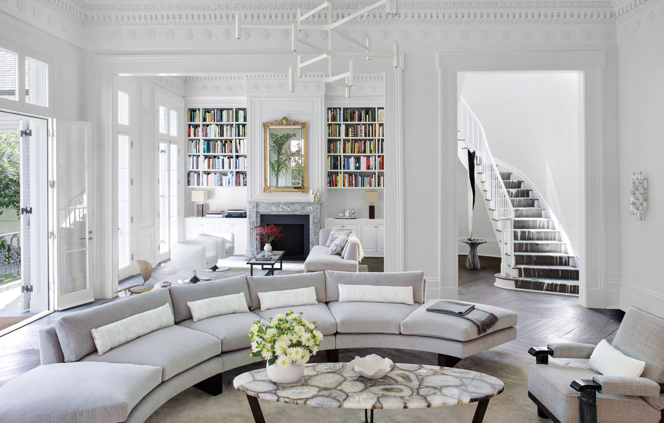 Elegant living room with curved sofa, coffee table, and staircase, featuring bookshelves, chandelier, and large windows.