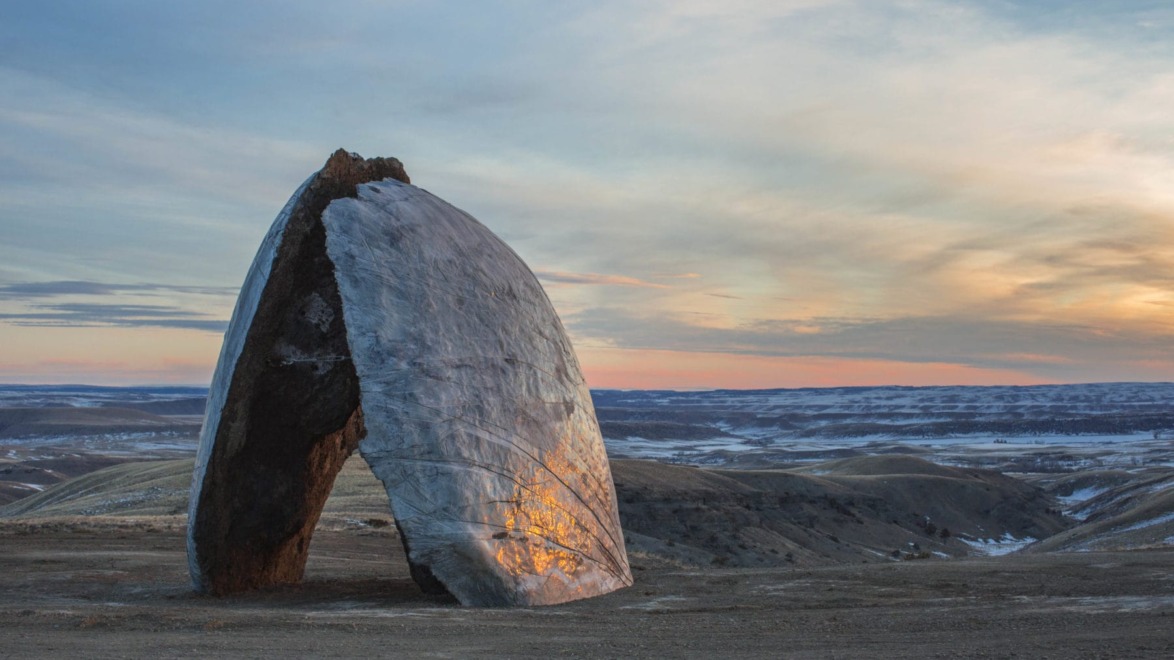 Large, arch-shaped rock sculpture standing on barren landscape with a colorful sunset sky in the background.