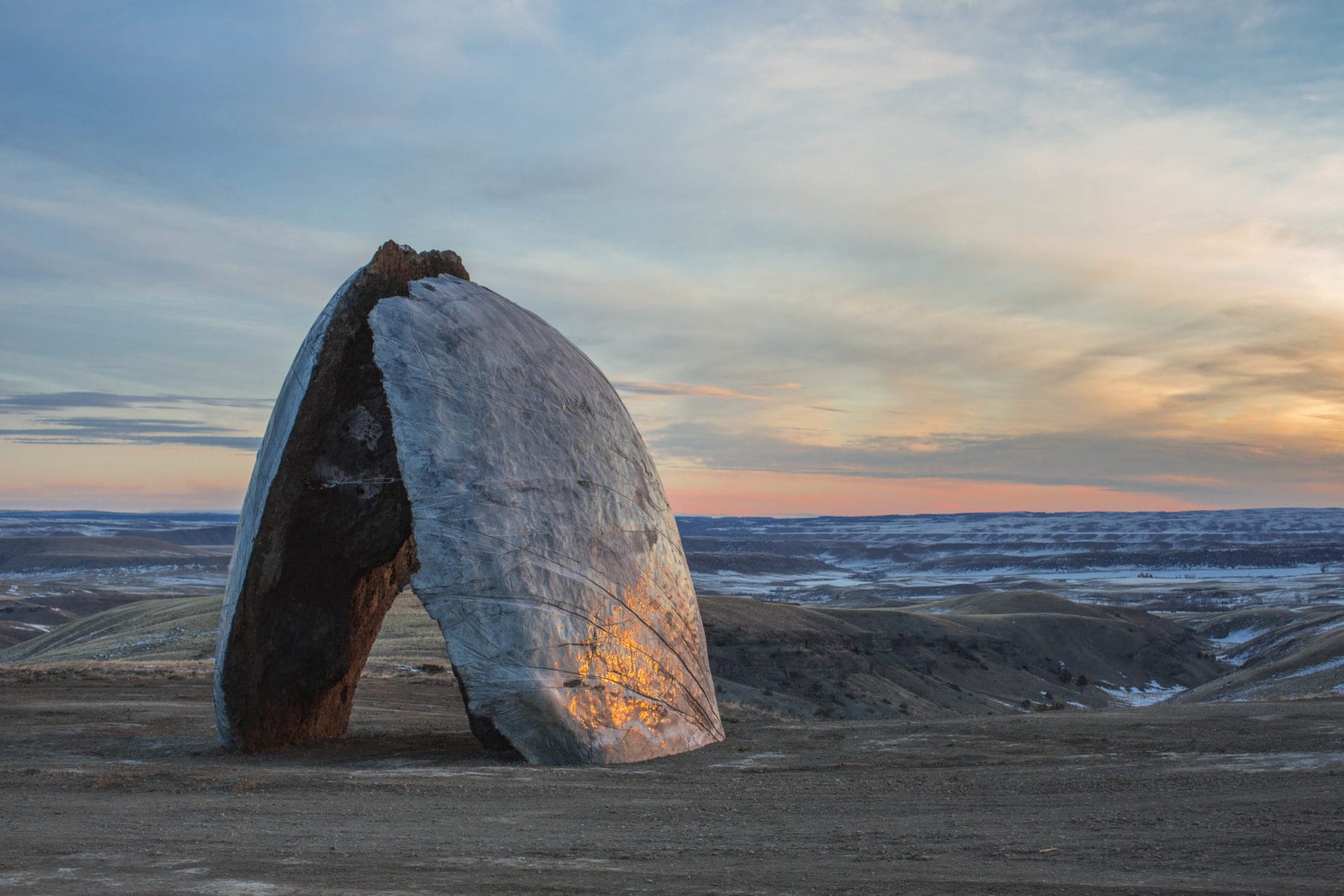 Large, arch-shaped rock sculpture standing on barren landscape with a colorful sunset sky in the background.