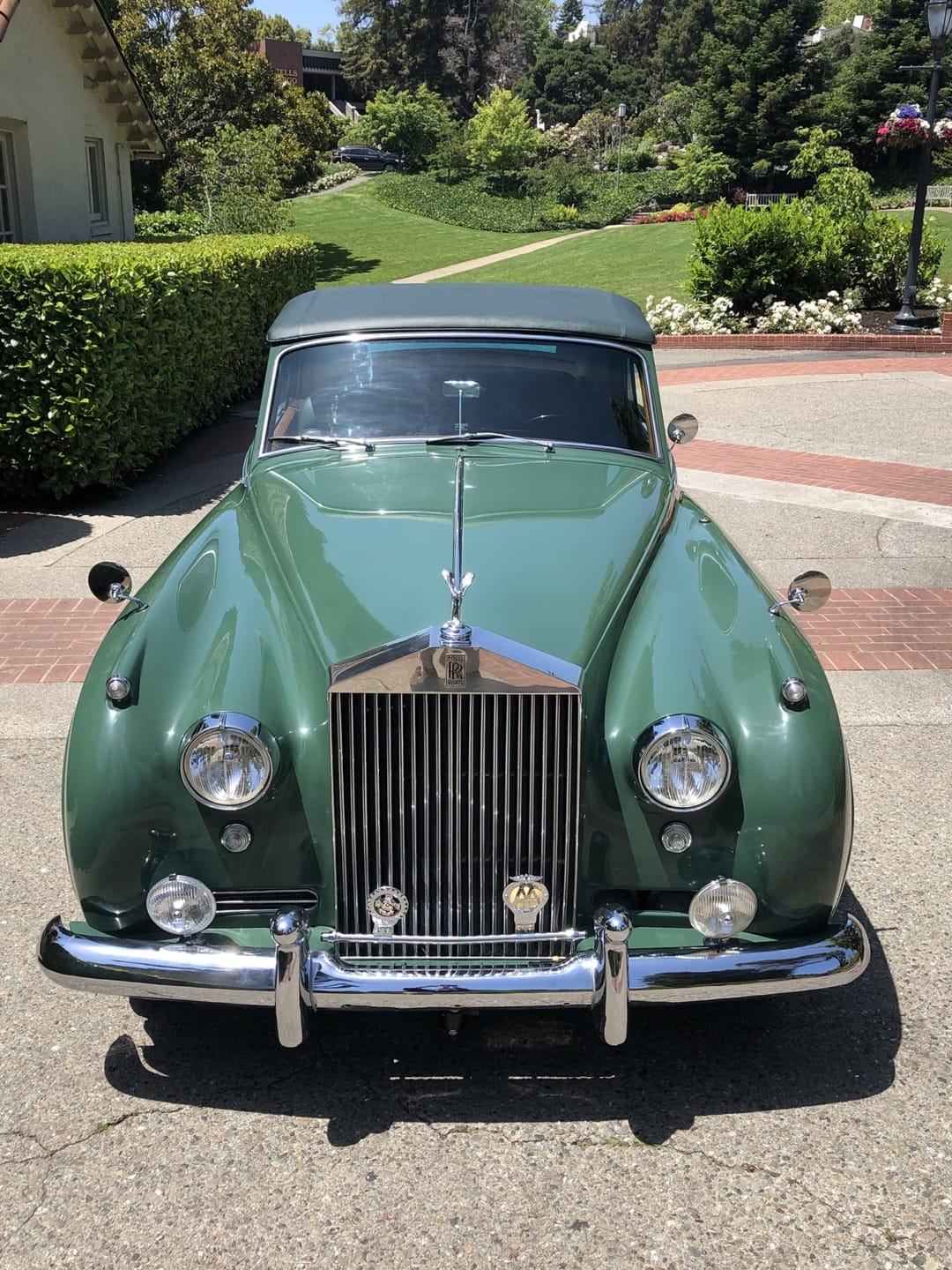 Green vintage car parked on a sunny driveway with lush greenery and a house in the background.