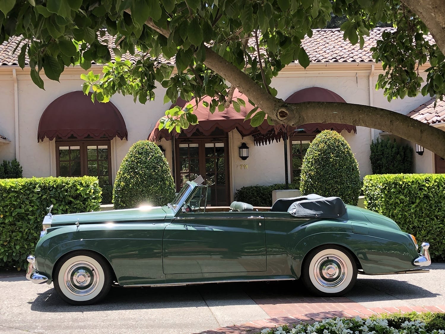 Classic green convertible parked in front of a house with red awnings and manicured shrubs.