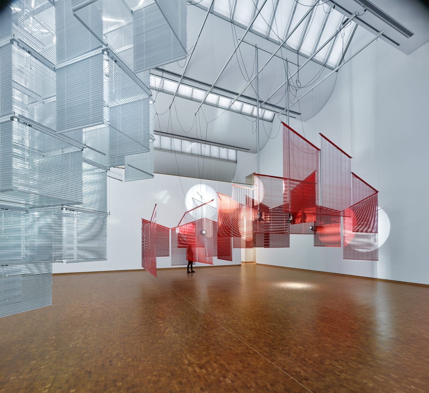Person observing a modern art installation with red and transparent panels suspended in a spacious gallery setting.