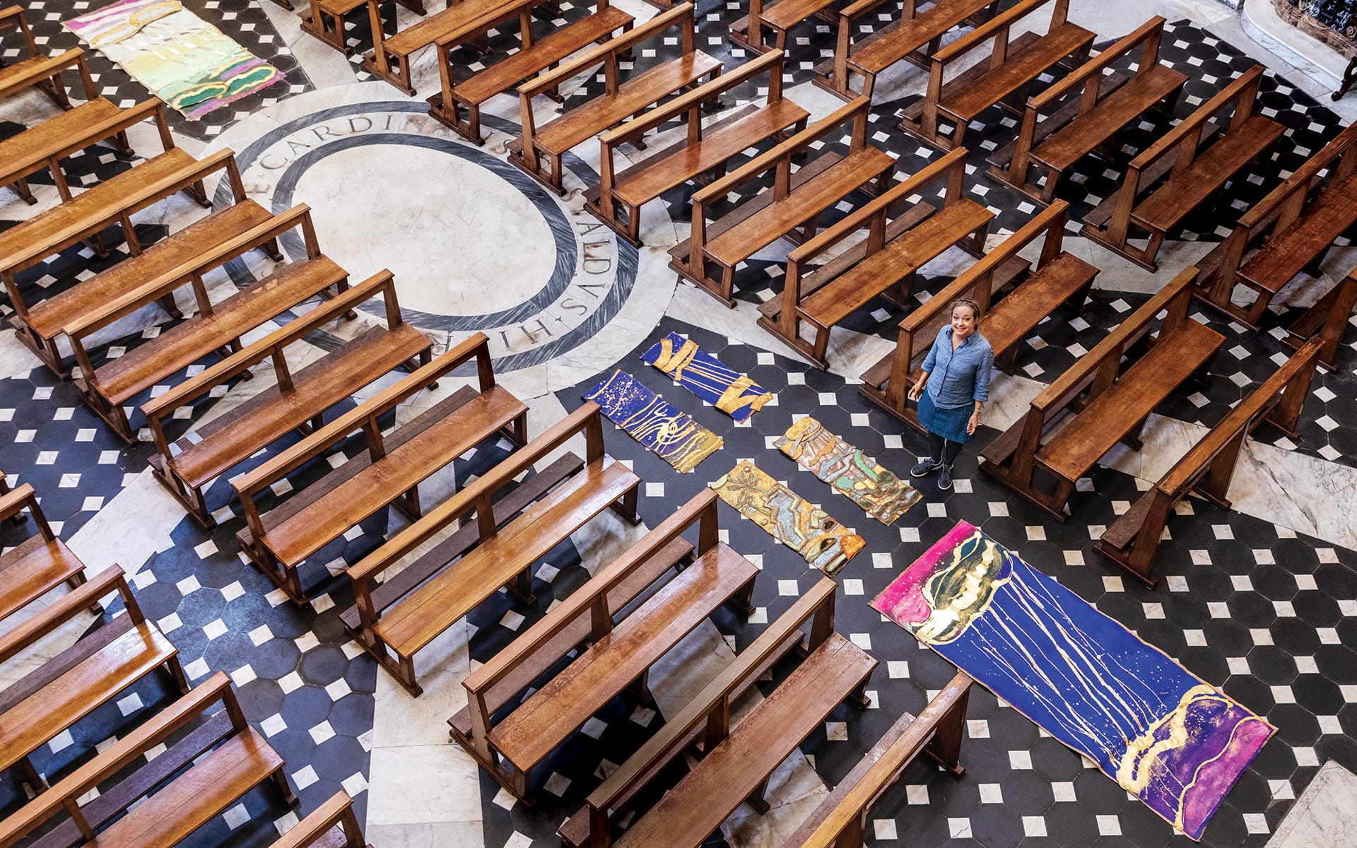 Person stands among artistic rugs on a checkered floor with rows of wooden pews in a spacious hall.