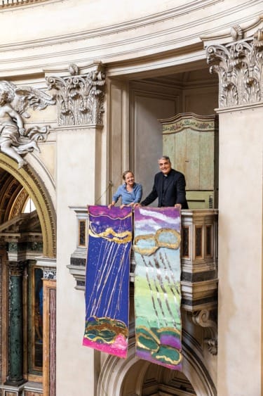 Two people standing on a balcony with colorful abstract banners in an ornate architectural setting.