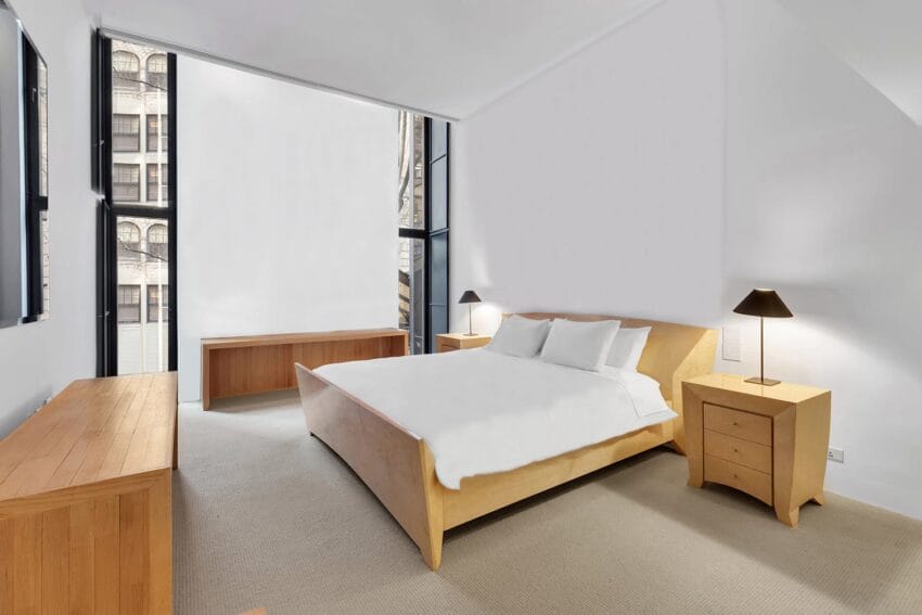 Modern bedroom with wooden furniture, large windows, and neutral decor. Room features a bed, nightstands, and a dresser.