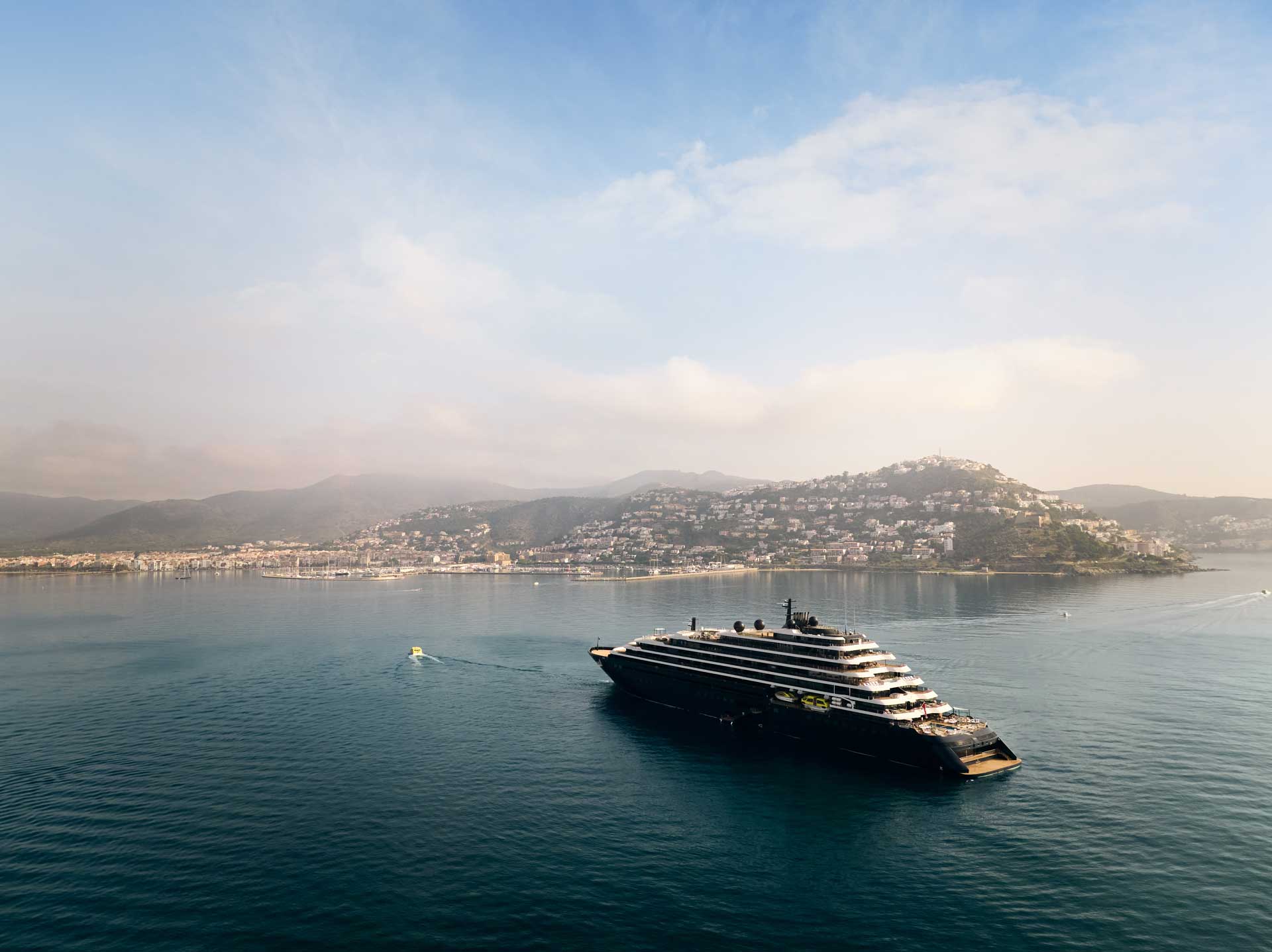 Luxurious yacht floating in calm blue waters with a mountainous coastline and misty sky in the background.