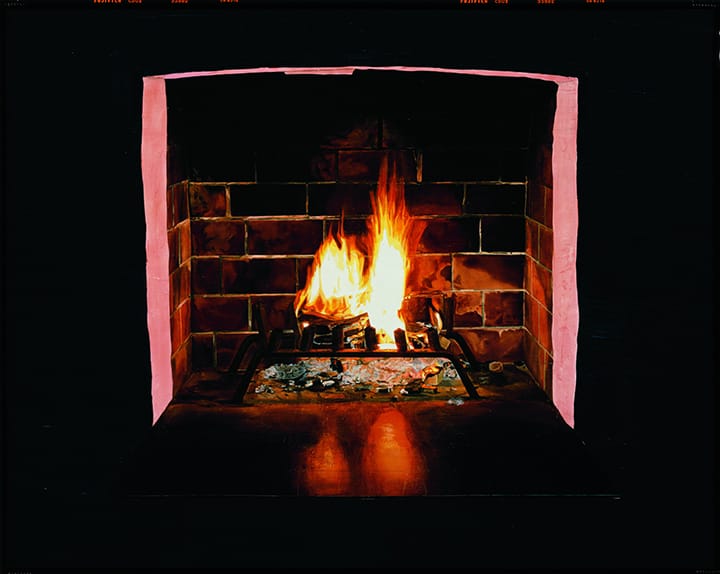 Lit fireplace with burning wood and glowing embers against a backdrop of dark, brick tiles.