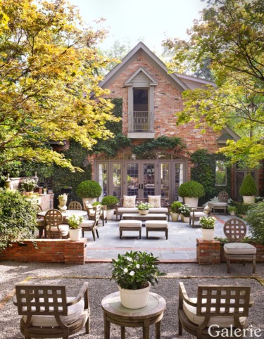 Charming brick house with lush garden, elegant outdoor seating, surrounded by vibrant greenery and small trees.