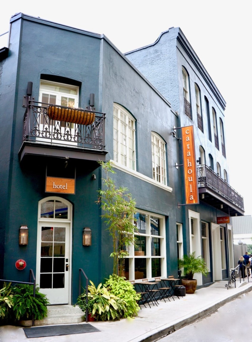 Historic urban hotel exterior with teal walls, metal balconies, and plants along the sidewalk on a bright day.