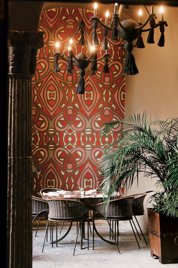 Stylish dining area with patterned wall, round table, wicker chairs, and chandeliers adorned with tassels in a tropical setting.