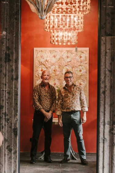 Two people standing in a stylish room with a colorful painting and a sophisticated chandelier overhead.