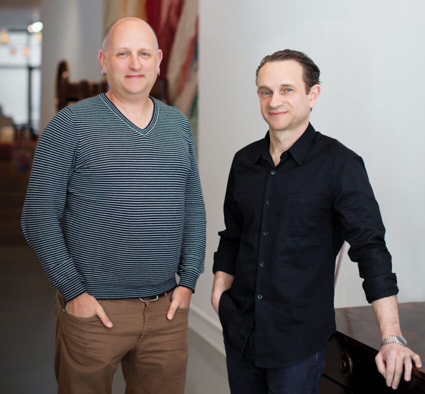 Two men standing in a modern office, one wearing a striped sweater and the other in a black shirt, both smiling.