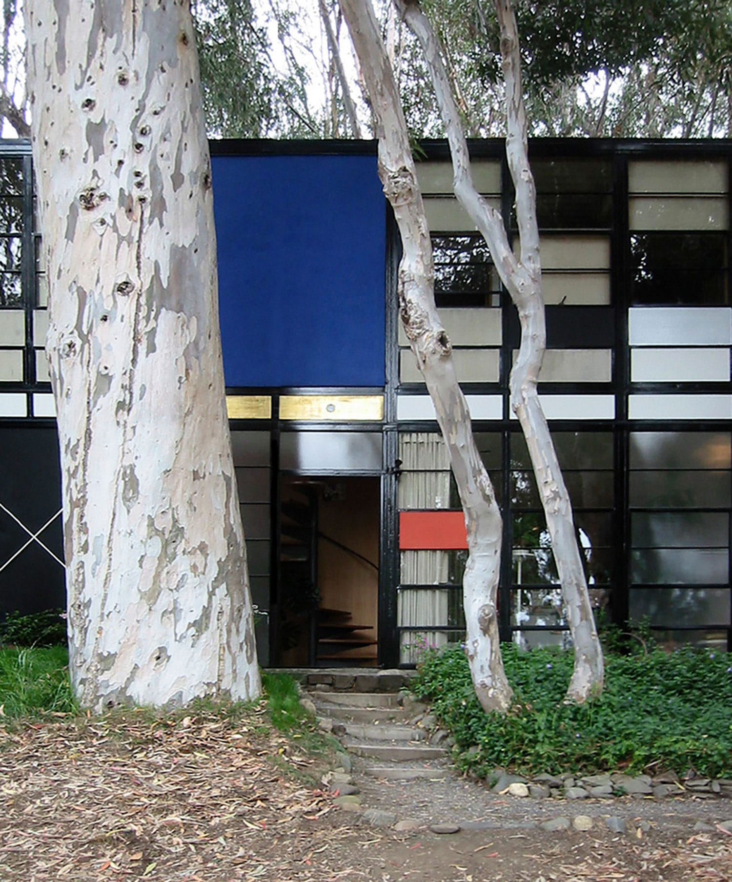 Exterior of a modernist house surrounded by tall trees, featuring a prominent blue panel and a pathway leading to the entrance.