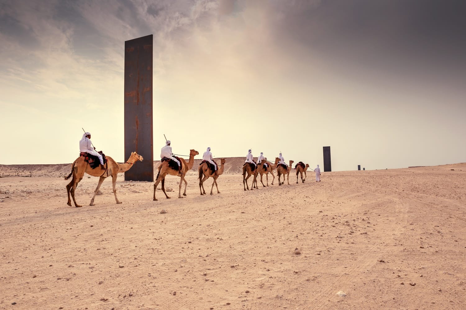 People riding camels in a desert landscape with towering abstract sculptures under a cloudy sky.