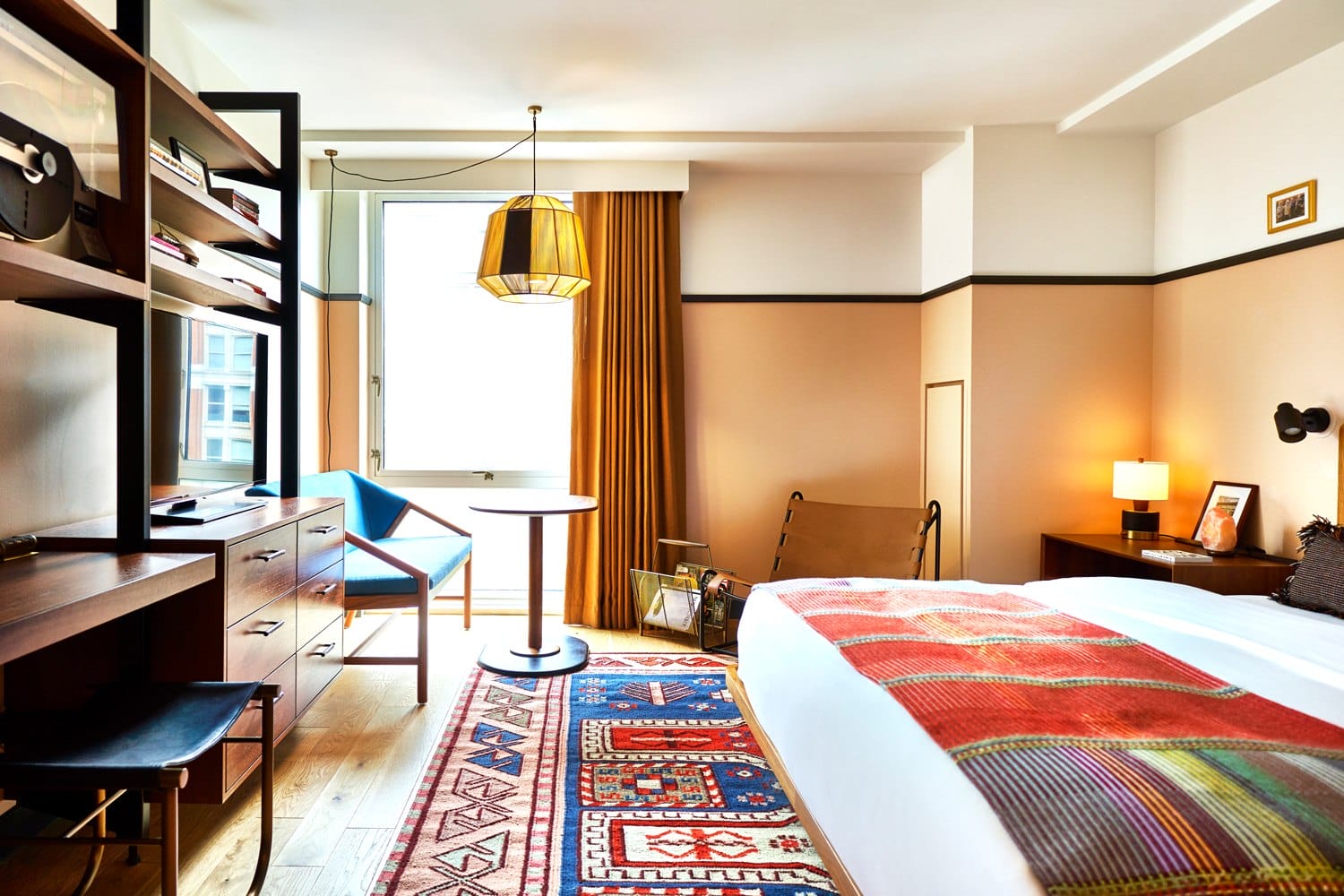 Modern hotel room with cozy bed, colorful rug, wooden furniture, and large window with golden curtains.