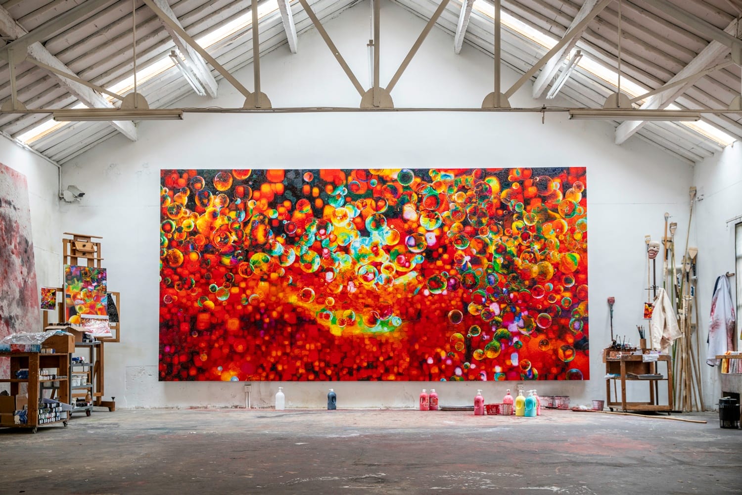 Large, colorful abstract painting with circular patterns in an artist's studio with high ceilings and scattered art supplies.