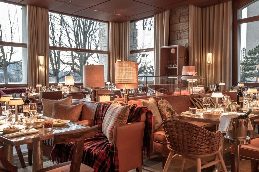 Cozy restaurant interior with wooden furniture, tables set with cutlery, plaid blankets, and large windows showing trees outside.