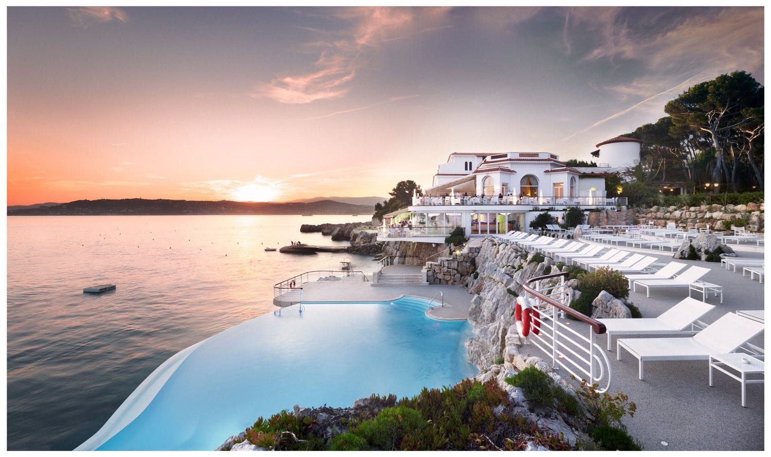 Luxury seaside resort with infinity pool at sunset, lounge chairs, and scenic ocean view.